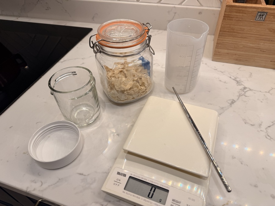 Equipment for rehydrating dried starter: small jar, flakes, measuring jug, scale, and stirrer
